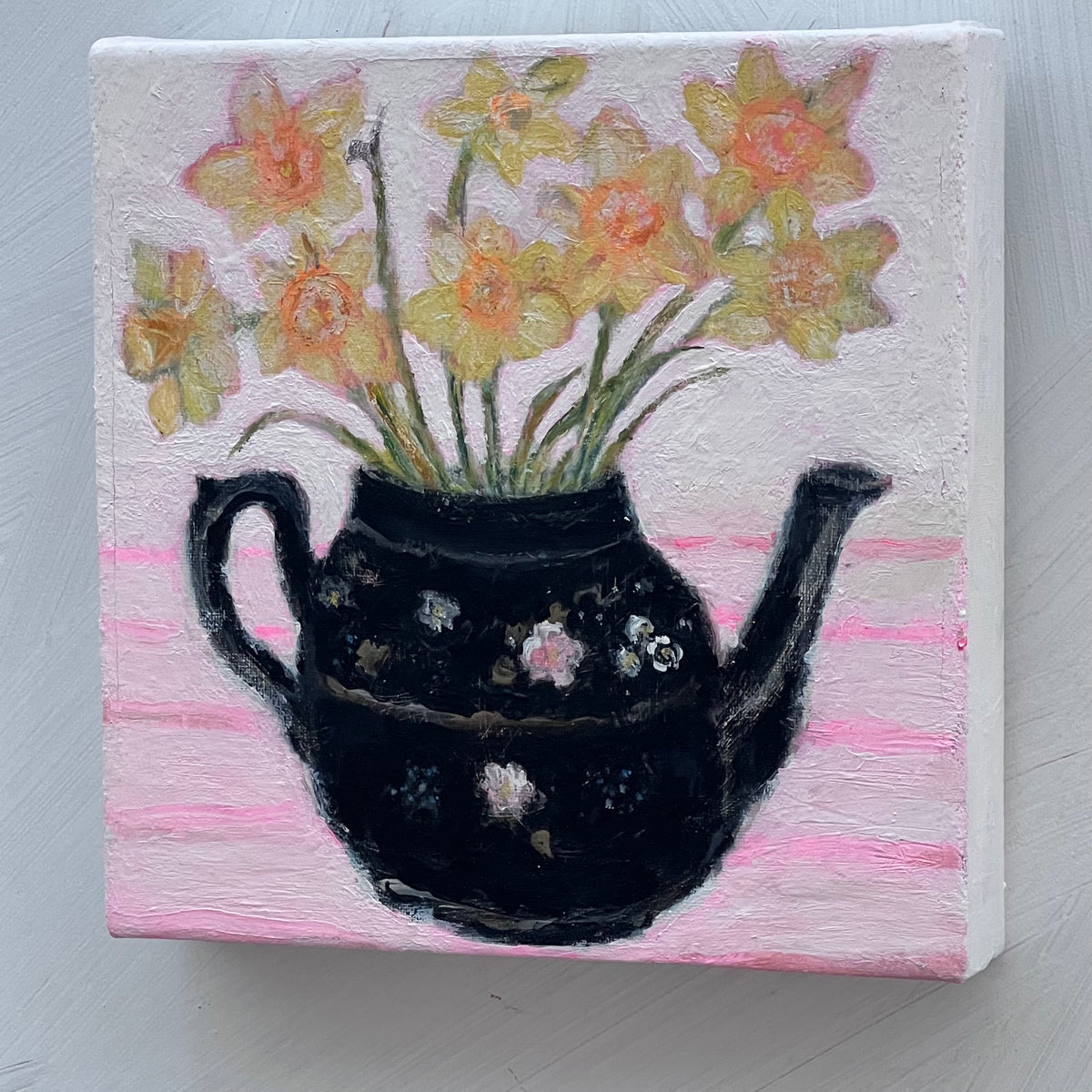 A painting of daffodils inside a black antique teapot on a canvas with a pink striped background.